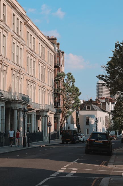 A wide street scene in Maida Vale featuring a row of cream-colored, multi-storey residential buildings with decorative window frames and iron balconies, situated on the left side of the image. The pavement in front is lined with trees and street lamps. Several cardboard boxes, wrapped in plastic and secured with packing straps, are positioned on the sidewalk near the building entrances, indicating ongoing packing or a recent move. A black van is parked partially on the pavement and partially on the road, suggesting loading or unloading activities. In the background, there are additional cars parked along the street, and a modern high-rise building can be seen against a blue sky with some scattered clouds. The scene reflects typical urban preparations for house removals and furniture transport, with visible packing materials and the presence of a vehicle ready for home relocation logistics. The environment appears calm, with a few pedestrians nearby, and the lighting indicates daytime with natural sunlight illuminating the surroundings.