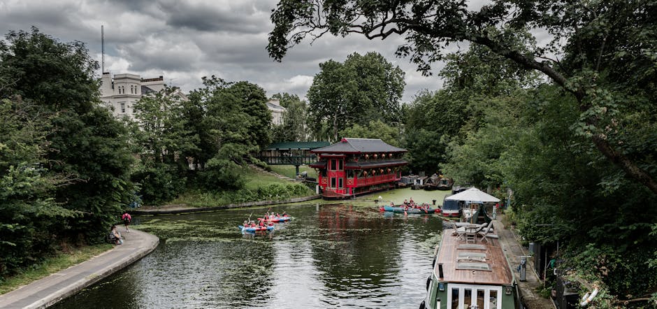 A scenic riverside scene showing several small kayaks and paddle boats floating on a calm waterway, with a red boathouse featuring traditional Asian architectural elements positioned on the opposite bank surrounded by lush green trees. On the near bank, a paved walkway runs alongside the river, with a person sitting on a bench and another walking nearby. To the right, part of a boat with a rusty metal roof and outdoor seating is docked, while the background features residential buildings partially obscured by dense foliage under a cloudy sky. This setting illustrates a peaceful outdoor leisure area, possibly near a park or city waterfront, with visible elements of boating and outdoor recreation as part of a home or community waterfront environment, relevant to relocation or moving services involving property and outdoor space management.
