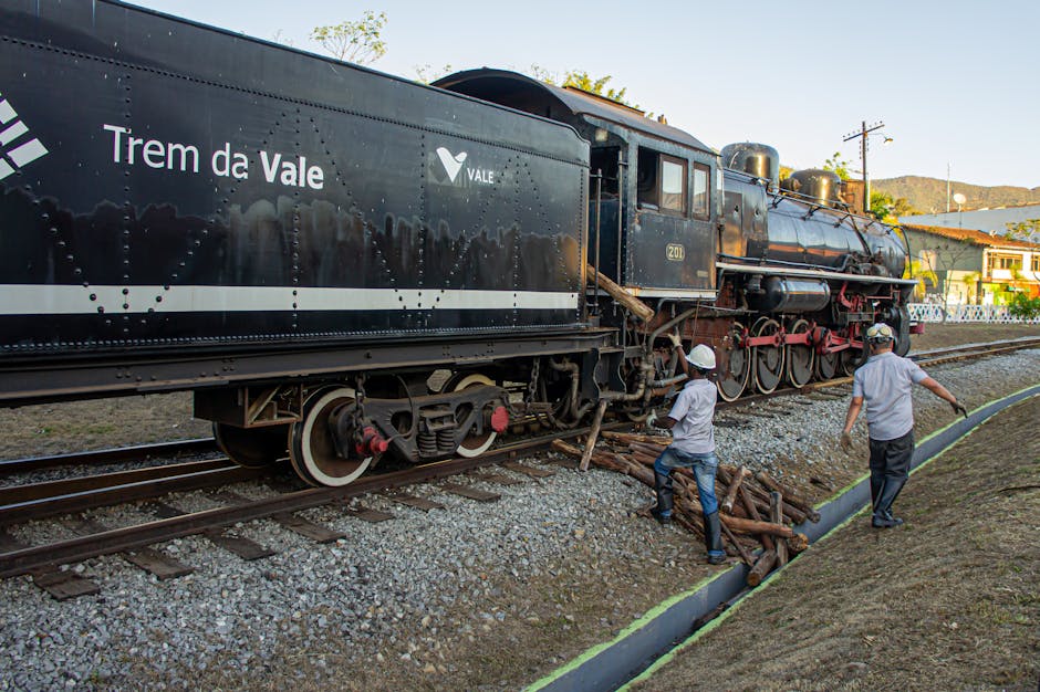 A vintage black steam locomotive labeled 'Trem da Vale' is positioned on railway tracks outdoors, with green trees and a residential area visible in the background under a clear sky. Two workers wearing white helmets and light-colored shirts are nearby; one is carrying a bundle of wooden logs, while the other appears to be walking along the path beside the track. The scene likely depicts a railway-related activity, such as rail maintenance or preparation for a moving operation involving a train. The environment suggests an outdoor setting connected to rail transport logistics. This image aligns with house removals or moving services by illustrating transportation and handling of large items or equipment, similar to furniture transport and loading processes that [COMPANY_NAME] offers for office or home relocations.