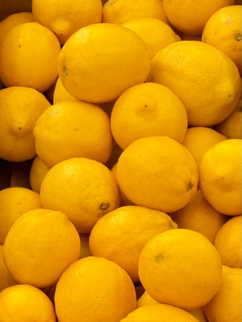 A close-up view of a large pile of fresh, bright yellow lemons with smooth, slightly textured skin. The lemons are piled together, occupying the entire frame, with some showing small blemishes or tiny dark spots. The vibrant yellow color contrasts against subtle shadows created by the natural lighting, highlighting the round shape and surface details of each fruit. There are no other objects or background elements visible, focusing solely on the collection of lemons which are typical produce items that might be found in a kitchen or fruit market. This image does not include any furniture, packaging, or moving-related items, and is unrelated to house removals or relocation services.