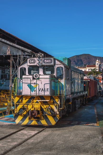 A vintage black steam locomotive labeled 'Trem da Vale' is positioned on railway tracks outdoors, with green trees and a residential area visible in the background under a clear sky. Two workers wearing white helmets and light-colored shirts are nearby; one is carrying a bundle of wooden logs, while the other appears to be walking along the path beside the track. The scene likely depicts a railway-related activity, such as rail maintenance or preparation for a moving operation involving a train. The environment suggests an outdoor setting connected to rail transport logistics. This image aligns with house removals or moving services by illustrating transportation and handling of large items or equipment, similar to furniture transport and loading processes that [COMPANY_NAME] offers for office or home relocations.