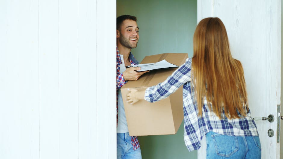 A man with dark hair and a beard, dressed in a plaid shirt over a white T-shirt, is standing inside a doorway holding a cardboard box with packing materials, while a woman with long red hair, wearing a blue and white plaid shirt and denim jeans, is outside the doorway reaching to pass a smaller cardboard box and some documents. They are engaged in a home relocation process, preparing for furniture transport as part of a house removals service. The scene takes place in a well-lit interior with a light-colored wall and white wooden door frame visible, and the setting suggests an organized loading process involving packing and moving tasks typical of professional removals conducted by [COMPANY_NAME], with objects positioned for an efficient furniture transport and packing and moving logistics.
