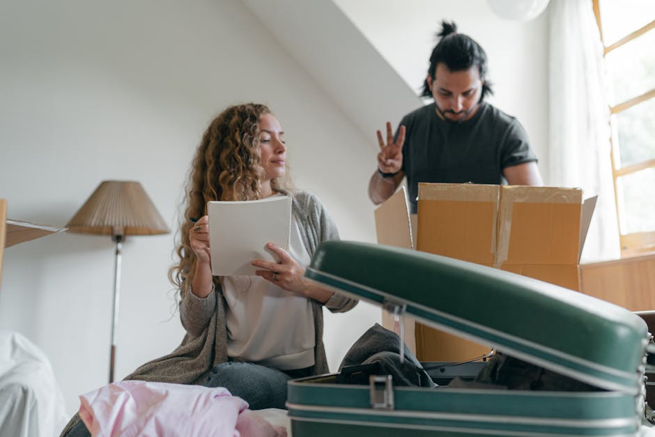 A woman with curly hair, wearing a beige cardigan and white top, is sitting on a bed in a room with natural light coming through a window with partially open blinds. She is holding an open notebook or folder and appears to be reviewing or organizing moving documents. Behind her, a man with dark hair, dressed in a dark t-shirt, is standing near a cardboard box and making a peace sign with his right hand while looking down at the box. In front of them, on the bed, there is a green and white suitcase that is open, with packing materials and clothing visible inside. To the left, a bedside lamp with a beige lampshade is on a wooden nightstand. The environment suggests they are in the midst of a home relocation process, surrounded by packing and moving supplies, with a focus on interior preparations for moving house. Man with Van Maida Vale occasionally supports the context of furniture transport and packing during a domestic move.