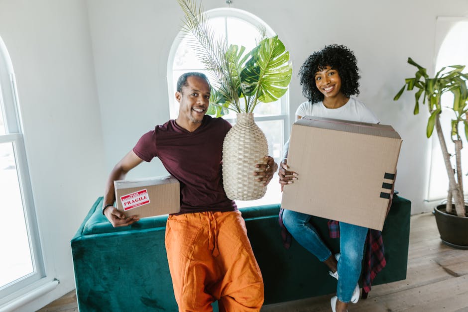 A man with dark hair and a beard, dressed in a plaid shirt over a white T-shirt, is standing inside a doorway holding a cardboard box with packing materials, while a woman with long red hair, wearing a blue and white plaid shirt and denim jeans, is outside the doorway reaching to pass a smaller cardboard box and some documents. They are engaged in a home relocation process, preparing for furniture transport as part of a house removals service. The scene takes place in a well-lit interior with a light-colored wall and white wooden door frame visible, and the setting suggests an organized loading process involving packing and moving tasks typical of professional removals conducted by [COMPANY_NAME], with objects positioned for an efficient furniture transport and packing and moving logistics.