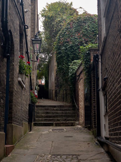 A narrow residential alleyway with stone steps leading up to a higher level, flanked by brick buildings on both sides. The left side features a black metal handrail, a wall-mounted lantern, and small flower pots with red and green plants. The right side has a white door partially visible at the bottom of the steps and a black drainpipe running along the wall. Dense foliage and creeping ivy cover the top of the wall at the end of the alley, with trees and a cloudy sky in the background. The concrete pavement shows signs of weathering, indicating an old urban environment. This setting illustrates the challenges faced during a home relocation or furniture transport through tight and uneven staircases, where careful handling and appropriate packing materials are necessary, as managed by professionals such as Man with Van Maida Vale.