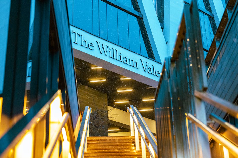 Exterior view of The William Vale building at dusk, showing illuminated entrance with modern stairs leading up to a glass door and the building's blue facade. The signage reads 'The William Vale' above the entrance, with metal railings along the staircase. The scene highlights the urban environment of a high-rise accommodation in Maida Vale, with lighting designed for evening visibility. Visible surroundings include dark reflections on the glass surfaces and part of the building's exterior walls. This setting exemplifies the type of location where professional removals services by Man with Van Maida Vale may be needed to facilitate furniture transport, home relocation, or packing and moving processes within a cityscape.