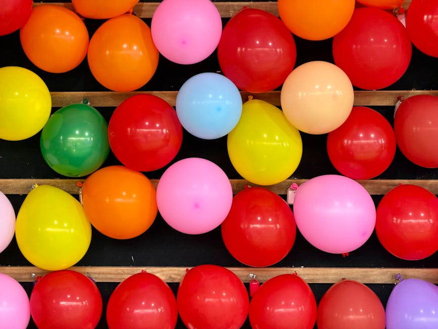 A collection of colorful balloons in shades of red, pink, yellow, orange, green, blue, and purple, arranged on wooden slats on a dark surface, with some balloons overlapping and others spaced apart. The balloons are inflated and glossy, reflecting light, and appear to be part of a celebration or decorative display. The background shows the balloons placed on a black or dark-colored surface, possibly at an event venue or a decorated indoor space. The image is related to house removals and packing processes, highlighting the importance of careful handling and transportation of delicate items such as balloons during a home relocation by Man with Van Maida Vale.