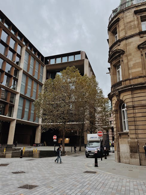 A narrow city street with tall, multi-storey buildings on both sides, showing a delivery van and a larger moving truck parked closely, partially obstructing the road. The delivery van is white with re