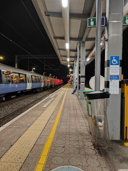 Nighttime view of Warwick Avenue Station platform with a row of stationary trains on the left side, illuminated by overhead lighting. The platform surface is paved with textured paving slabs, featuring a tactile yellow line near the edge for safety. On the right side, there is a covered waiting area with a metal canopy supported by vertical poles. Attached to one pole is a blue sign indicating accessible facilities for disabled passengers. A metal and plastic waste bin is positioned beneath the sign. The station appears empty, with no passengers visible, and the surrounding environment is dark beyond the platform lighting. This scene captures the typical layout of a London suburban station, with clear spaces for passengers to wait and prepare for home relocation or furniture transport, as part of the moving or packing process, which companies like Man with Van Maida Vale often assist with.