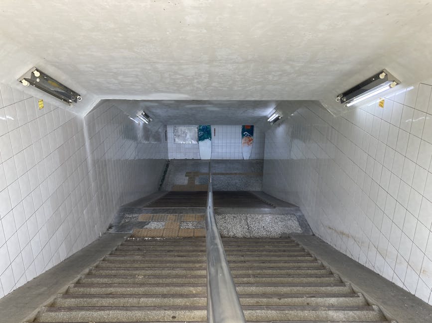 The image depicts a downward view of a short staircase leading from street level to the underground entrance of Warwick Avenue Station, with white tiled walls on either side. The stairs are made of concrete or stone with visible wear and a metal handrail running down the center. On the left and right walls, there are fluorescent light fixtures providing illumination. At the bottom of the stairs, the station platform is visible, featuring advertisements or posters attached to the tiled wall, along with a small section of tiled flooring and part of the station's entrance door. The environment appears clean and well-lit, with no people present, illustrating a typical access point for public transportation that could be used during home relocation or moving processes involving transport to or from Maida Vale, as handled by Man with Van Maida Vale.